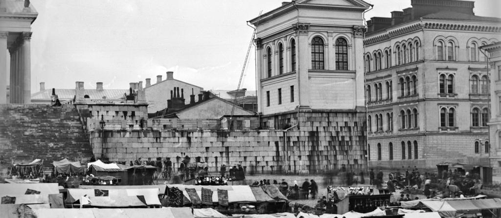 Senaatintori on täynnä markkinakojuja ja ihmisvilinää. Etualalla näkyy mm. rinkelikauppiaita myyntipöytiensä ääressä. Taustalla nousee Tuomiokirkon oikeanpuoleinen tasanne.