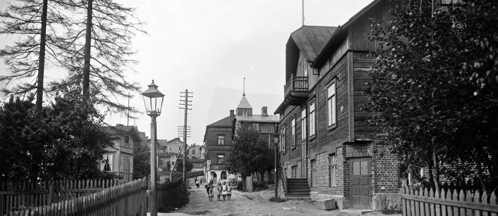 Katunäkymä Länsi-Pasilassa vuonna 1912. Kuvassa on työväen asuntoja suurempi puuhuvila. Hiekkatiellä kävelee muutama ihminen.