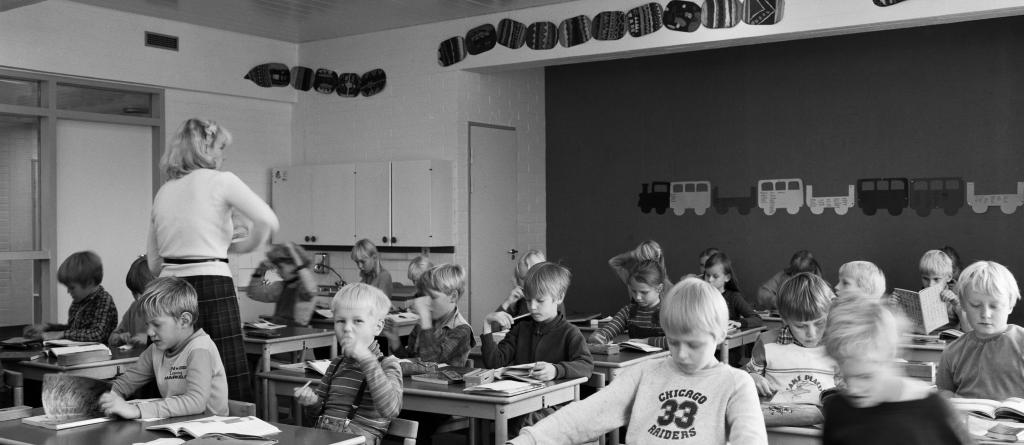 Alakoululaisia oppitunnilla luokassaan. Oppilaiden pöydillä on kirjapinoja, opettaja kiertää luokassa.