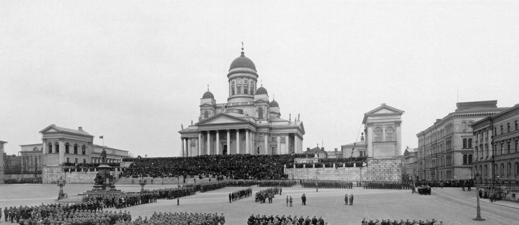 Jääkärit ovat järjestäytyneet paraatimuodostelmiin ympäri Senaatintoria. Ryhmittymien keskelle on kokoontunut oletettavasti jääkäriprikaatin päällystöä. Yleisö on kerääntynyt seuraamaan tapahtumia tuomiokirkon portaille.