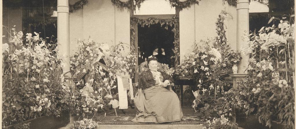 Aurora Karamzin fotograferad på sin 90-årsdag sittande framför ytterdörren till sin villa, omringad av blomplanteringar. En liten ljus hund sitter framför henne på trappan. 