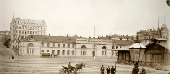 Hevosajureita rattaineen ja herrasväkeä Eteläsatamassa Pakkahuoneentorilla. Taustalla entinen sotaväen makasiini, vuodesta 1888 pakkahuone, Läntinen Rantakatu 10 (nyk. Eteläranta 10). Kadulla hevosvetoinen raitiotievaunu. Makasiinin takana kohoaa 1960-luvun lopulla purettu viisikerroksinen kivitalo osoitteessa Unioninkatu 14 - Eteläinen Makasiinikatu 3.