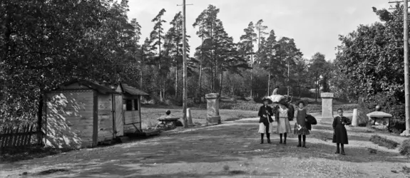 Kaupungin raja, Vanha tulliportti. Läntinen Viertotie 80 (nykyinen Mannerheimintie 108). Neljä lasta seisoo keskellä tietä, taustalla näkyy hevoskärryä ajava mies. Tien vasemmalla puolella on matala puumökki, jonka takana tien kummallakin puolella on kivipaadet, joiden päällä tullinpuomi on aikoinaan levännyt.