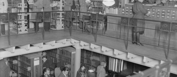 En vy mot två våningar i biblioteket på Rikhardsgatan. På den övre våningen studerar kunderna böcker och ett kartotek, på den nedre köar de till betjäningsdisken. 