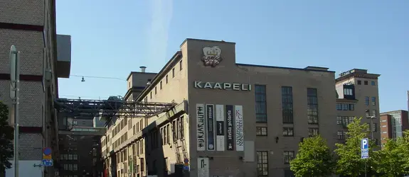 Kaapelitehdas kuvattuna parkkipaikan puolelta kesäaikaan. Julkisivuun on kiinnitetty valotaulu KAAPELI ja Radio City sekä ripustettu sivuttain plakaatteja, joissa lukee esimerkiksi HURJARUUTH ja ZODIAK.