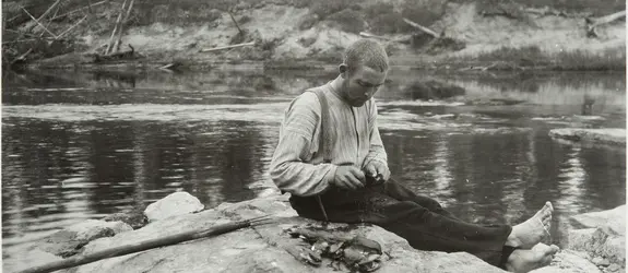 En man sitter på en klippa vid en å och öppnar musslor med en kniv. Bredvid honom är en hög öppnade musslor.