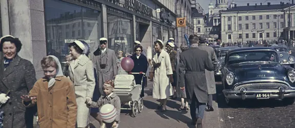 Människor med studentmössor promenerar på gatan. Södra kajen 18. I bildens högra kan en personbil av märket Buick och årsmodell 1954.