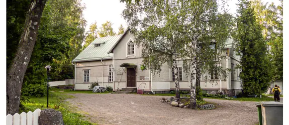 Puurakenteisen talon edessä kasvaa kolme koivua. Oven pielessä oikealla on lumikola, ovesta vasemmalla puutarhapöytä ja -tuoleja.
