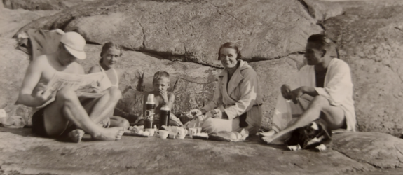 Två män och kvinnor med en pojke i mitten sitter på strandklippor med matsäck. Den ena mannen bär keps och läser en tidning. 
