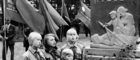 Neljä nuorta pioneeriasuista tyttöä laulaa Santahaminan veljeshaudalla punaisten muistomerkin edustalla. Taustalla lippuja piteleviä miehiä.
