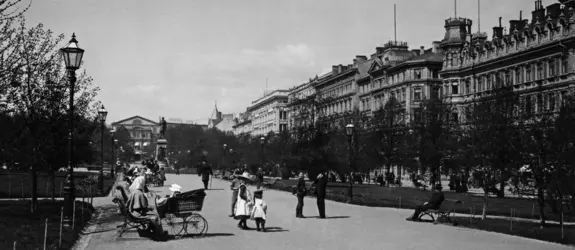 På Runebergsesplanaden finns människor som sitter på bänkar och promenerar. Till höger syns Norra Esplanadens höga byggnader.