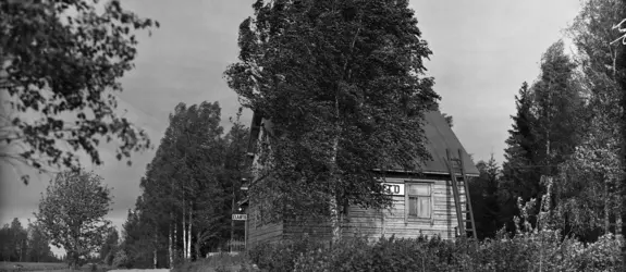 Vasemmalla on kapea hiekkatie, oikealla puolitoistakerroksinen puurakennus, jonka ympärillä on koivuja. Rakennuksen seinässä lukee ELANTO.
