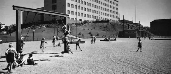 Barn på Brahe lekplats i Berghäll. I förgrunden leker en grupp barn i gungor. I bakgrunden på en backe höjer sig Aleksis Kivis folkskola.