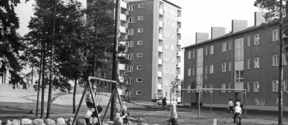Etualalla leikkipuistossa leikkiviä lapsia. Osa lapsista on hiekkalaatikolla, muutama kiikkuu ja parilla on polkupyörät. Leikkipuisto on kivistä kasatun aidan sisälle jäävä hiekkakenttä. Taustalla näkyy kerrostaloja ja puita.