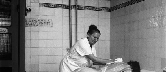 A washerwoman dressed in a white coat soaping up the back of a man lying face down. There are white tiles and a sign with the text “Using soap in the sauna or going to the sauna covered in soap is strictly prohibited” in the background.