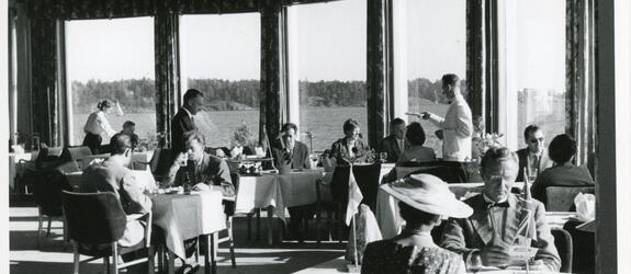 Brändö kasino, Silversundsvägen 49, restaurangsalen och kunder. Serveringspersonal i bakgrunden. Genom fönstren skymtar en havsutsikt.