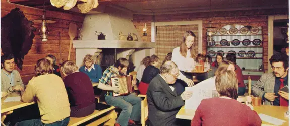 Pirtin tuvan tyyliin sisustettu ravintolatila, jossa ihmiset istuvat esimerkiksi lukemassa lehtiä. Mies soittaa haitaria, tarjoilija on tuomassa olutta ja limonadia tarjottimella. 