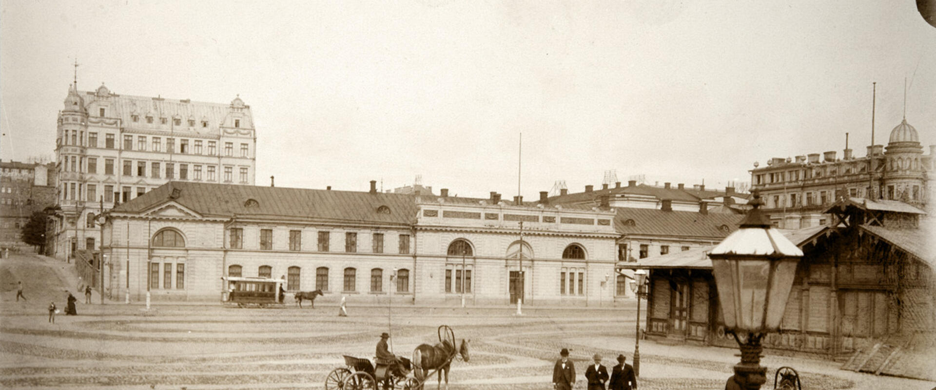 Hyrvagnar med sina hästar och herrskap vid Södra hamnen på Packhustorget. I bakgrunden det tidigare arméförrådet, från 1888 packhuset, Västra Hamngatan 10 (nuvarande Södra kajen 10). På gatan en hästdragen spårvagn. Bakom förrådet syns ett femvåningshus från 1960-talet vid hörnet av Unionsgatan 14 – Södra Magasinsgatan 3.