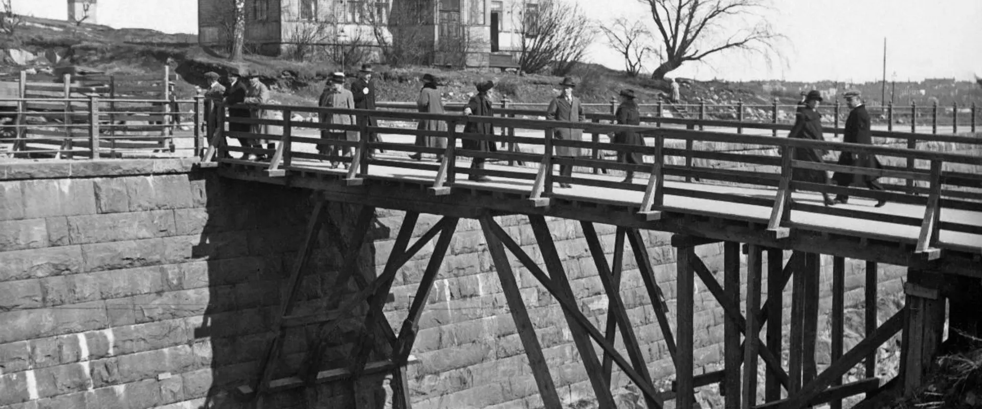 Människor promenerar på en träbro. Under bron löper hamnbanan. På andra sidan bron står en trävilla med en våning samt några kala björkar. I bildens vänstra kant syns Nationalmuseets torn.