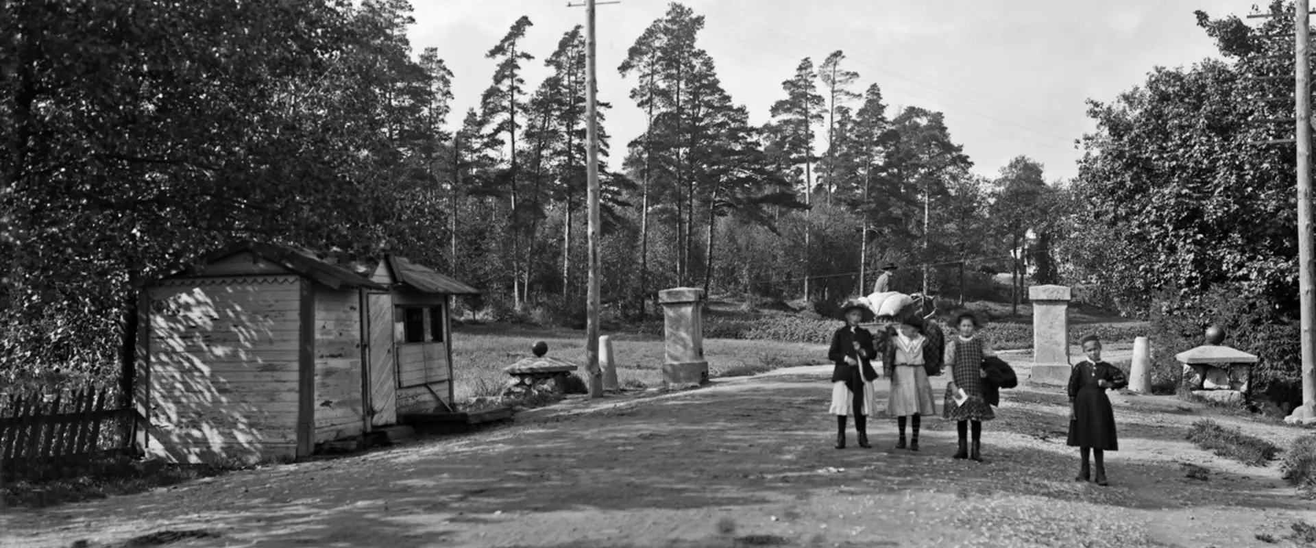 Stadsgräns, gammal tullport. Västra Chaussén 80 (numera Mannerheimvägen 108). Fyra barn står mitt på vägen, i bakgrunden syns en man som kör med häst. Till vänster om vägen finns en låg trästuga, och bakom den på båda sidorna om vägen finns stenpelare, som en tullbom tidigare vilat på.
