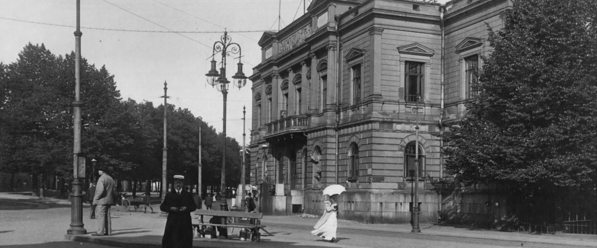 Framför Gamla studenthuset står en dekorativ gaslykta. Längs gatan går en kvinna i lång vit klänning och med ett vitt parasoll, och en man i studentmössa.