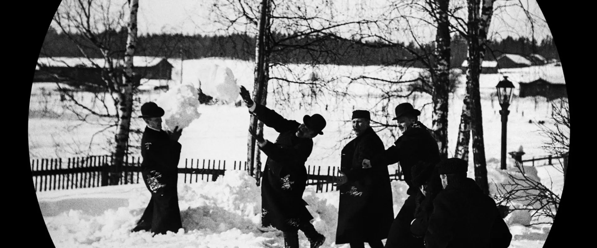 Män klädda i hattar och långa kappor leker med snöbollar. Bakom dem finns björkar och en gaslykta.