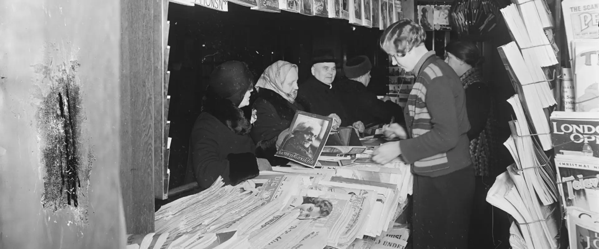 Bilden har tagits inne i en tidningskiosk och på den syns ett stort antal tidningar på olika språk samt försäljaren i sidoprofil. Vid disken finns fyra kunder i vinterkläder.