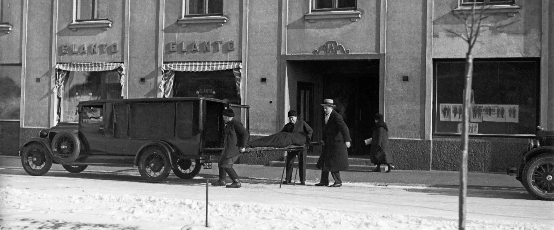 En snötäckt gata och trottoar framför ett höghus. Framför huset står en ambulans och annan bil. Tre män håller på att lyfta en en bår i ambulansens bakdel. I bakgrunden syns en kvinna som går förbi.