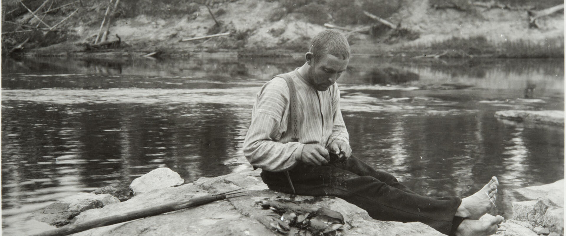 En man sitter på en klippa vid en å och öppnar musslor med en kniv. Bredvid honom är en hög öppnade musslor.