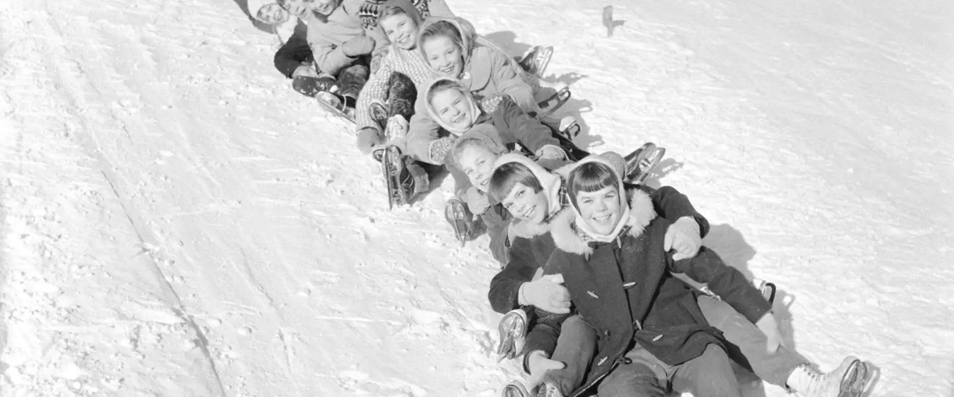 Yli kymmenen lasta on muodostanut pitkän letkan, joka laskee yhdessä pyllymäkeä. Ainakin etummaisilla on luistimet jalassa. 
