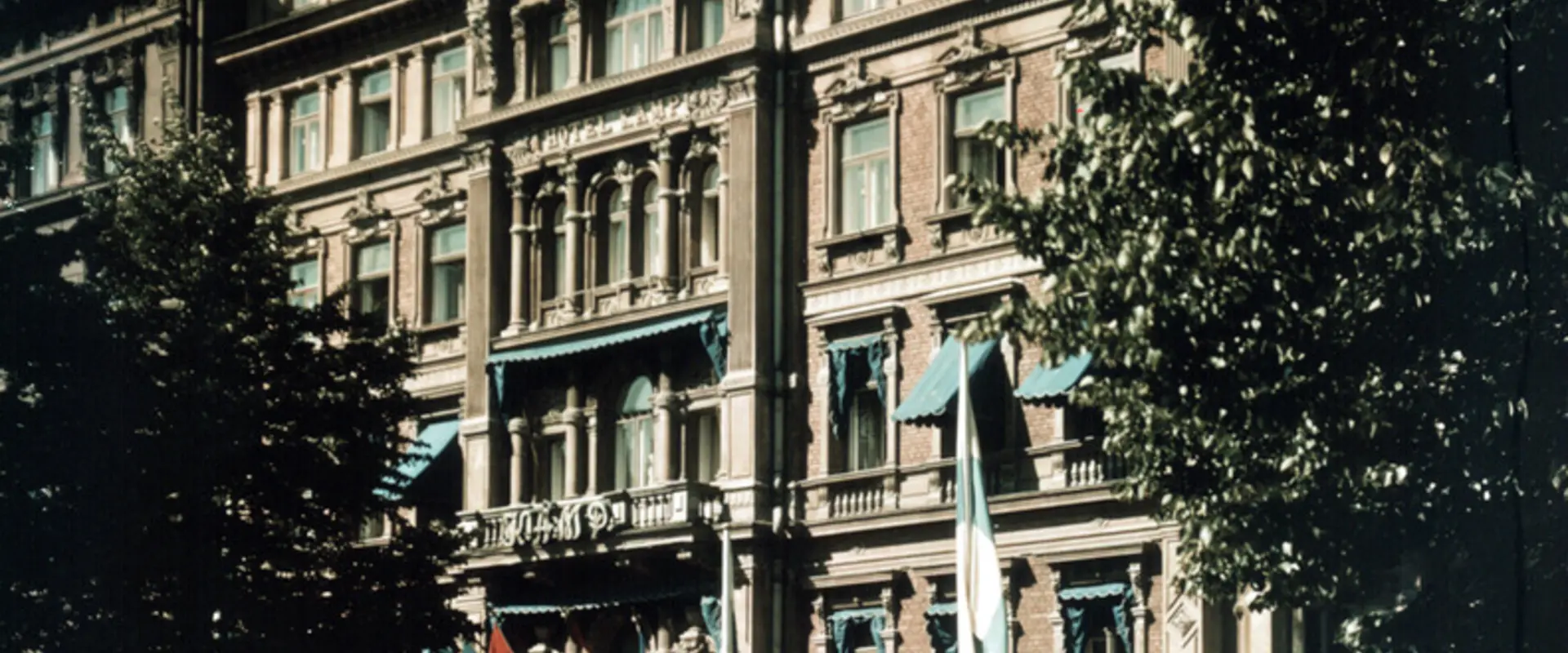 Hotell Kämps fasad fotograferad på sommaren. På Esplanaden syns en vit bil, i förgrunden står fyra kvinnor.