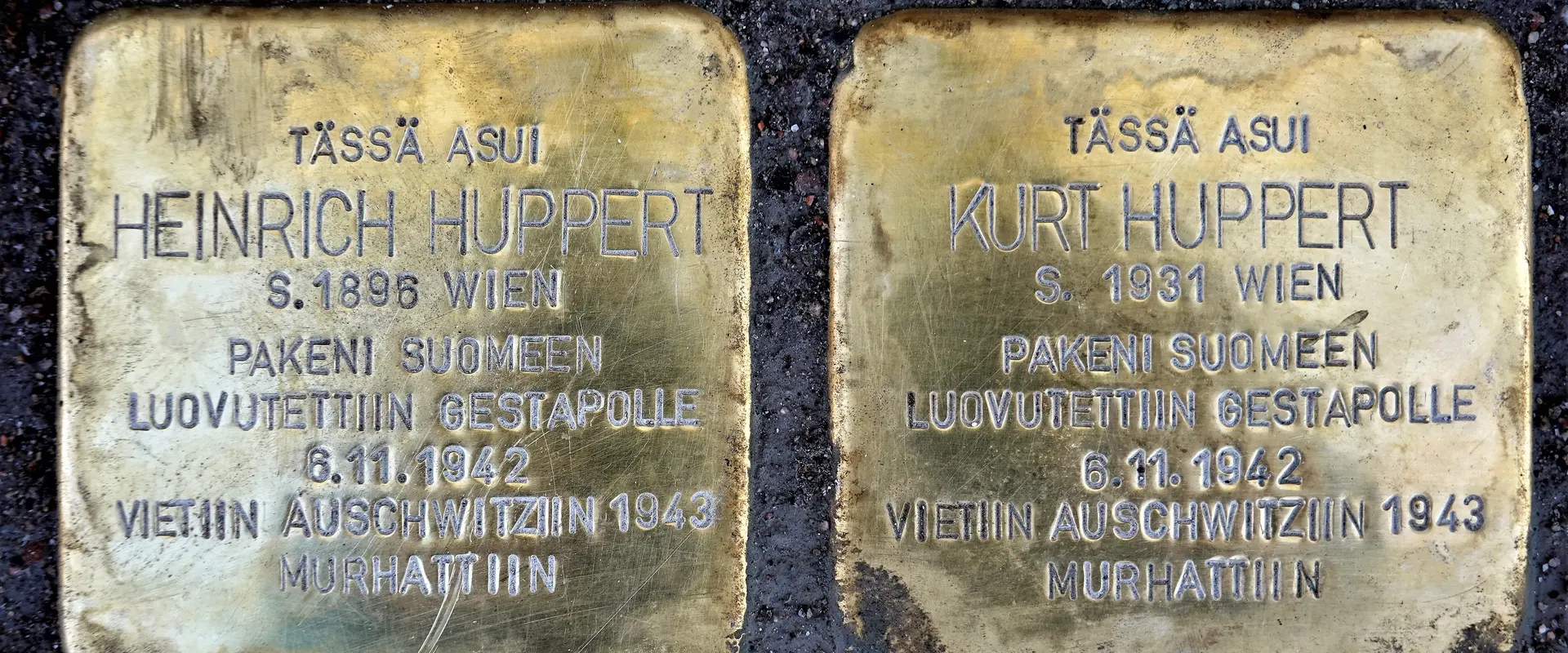 Två metallplattor i asfalten. På dem finns texterna: Här bodde Heinrich Huppert född 1896 Wien Flydde till Finland Utlämnades till Gestapo 6.11.1942 Fördes till Auschwitz 1943 Mördades