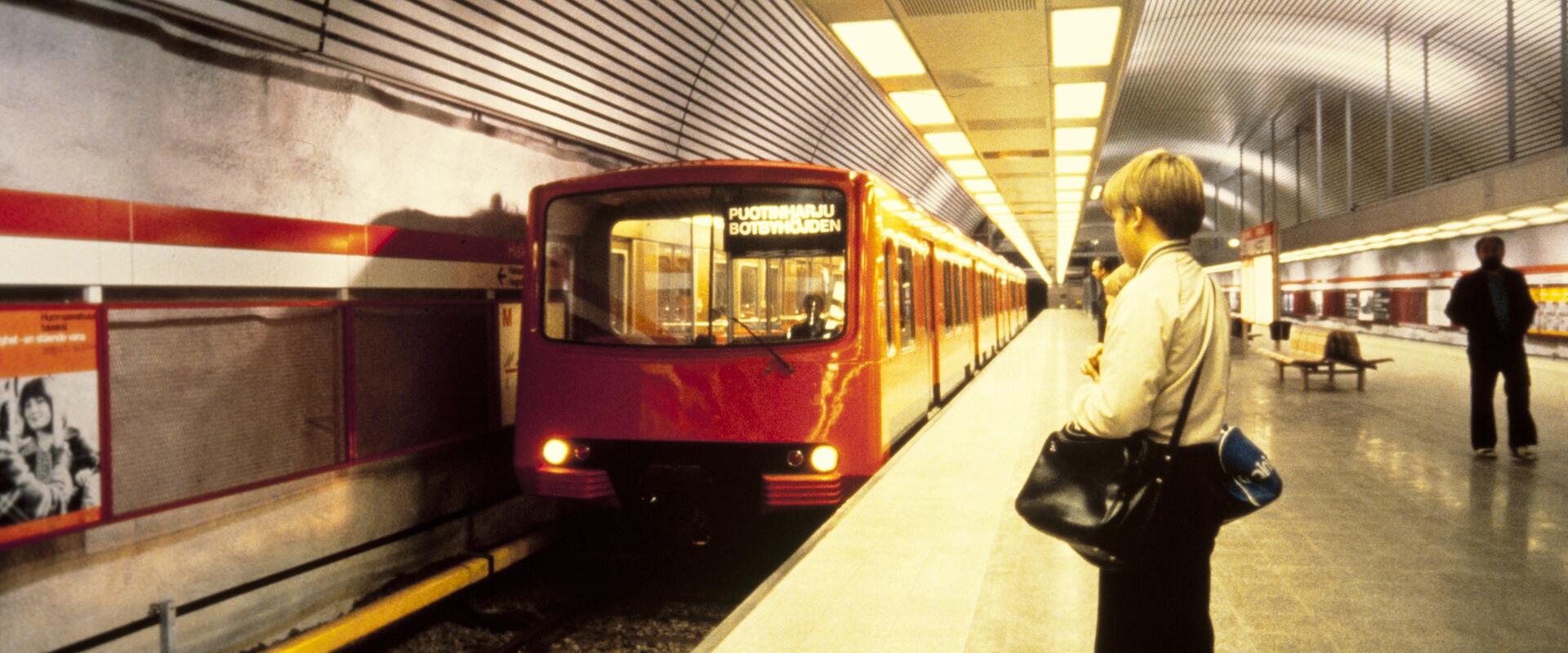 Metrotågen anländer till stationen. I förgrunden väntar en ung man i ylletröja med två väskor.