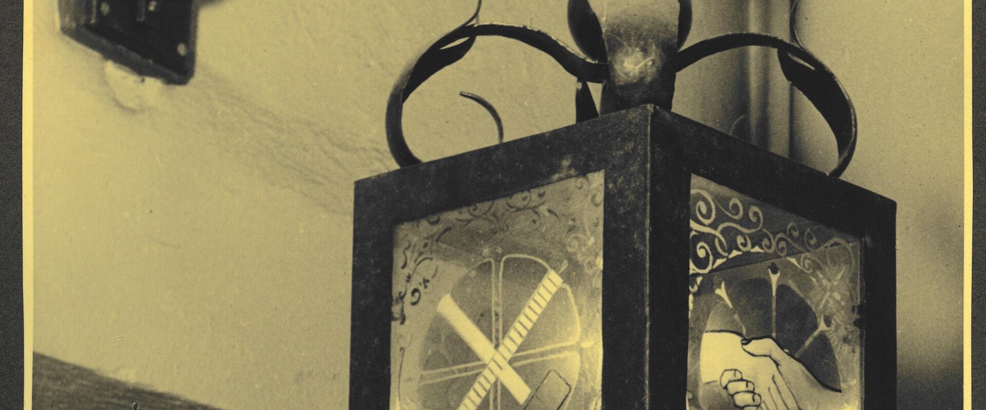 En rektangulär lykta som hänger ovanför en dörr. Lyktan har graverade glasväggar som har dekorerats med frimurarsymboler som en hammare och en måttstock och två händer som håller i varandra.