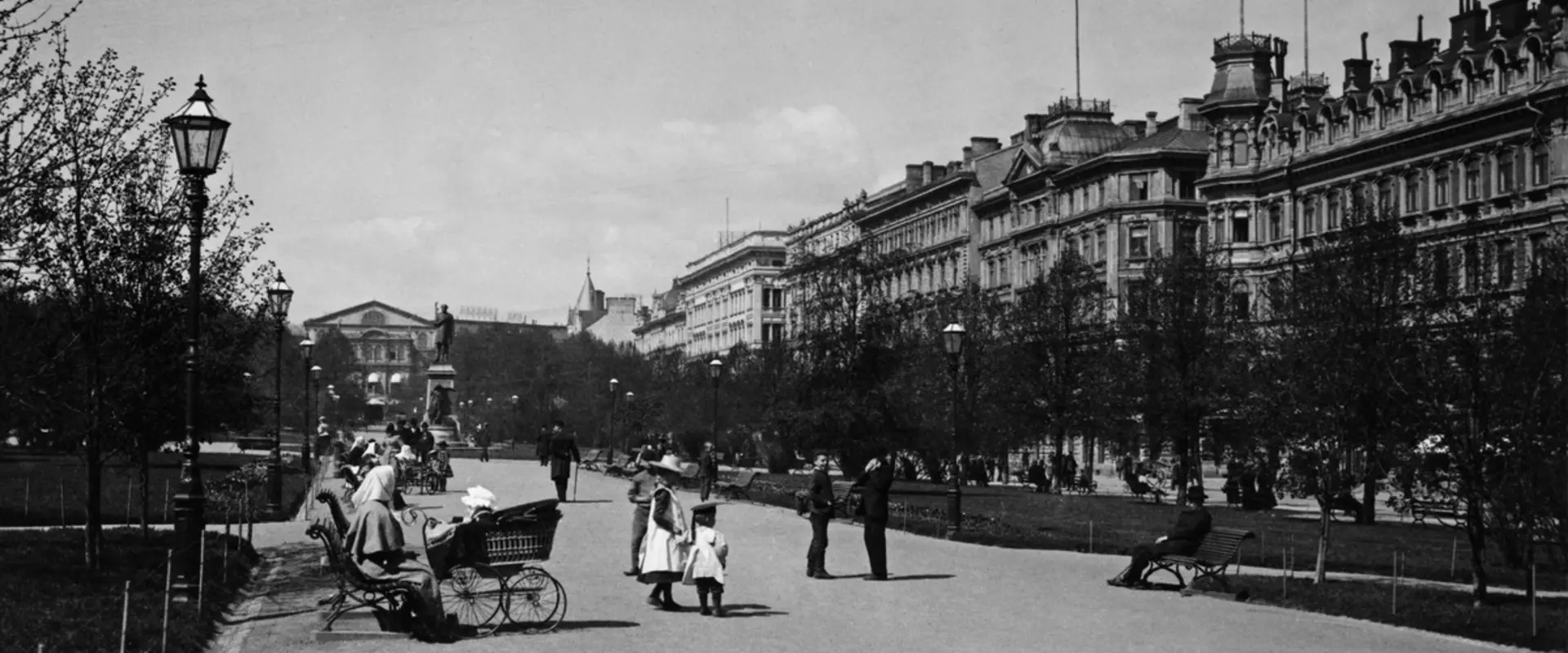 På Runebergsesplanaden finns människor som sitter på bänkar och promenerar. Till höger syns Norra Esplanadens höga byggnader.