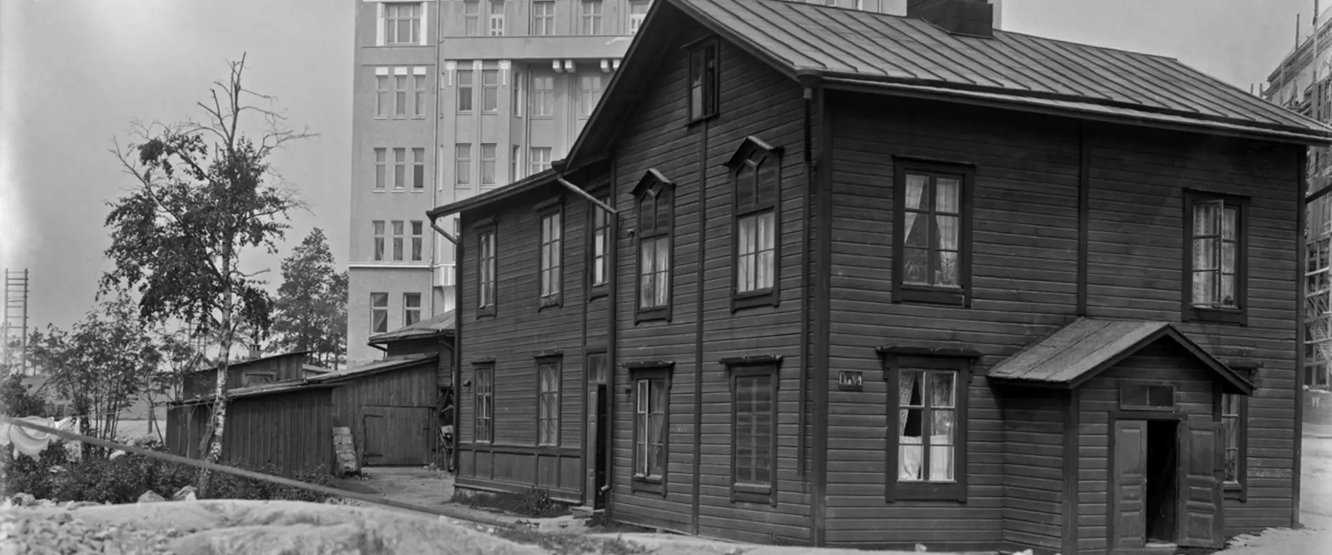Kuvan keskellä on kaksikerroksinen puurakennus, jonka vasemmalla puolella on matalia piharakennuksia. Taustalla kohoaa samana vuonna valmistunut kuusikerroksinen kivitalo. 