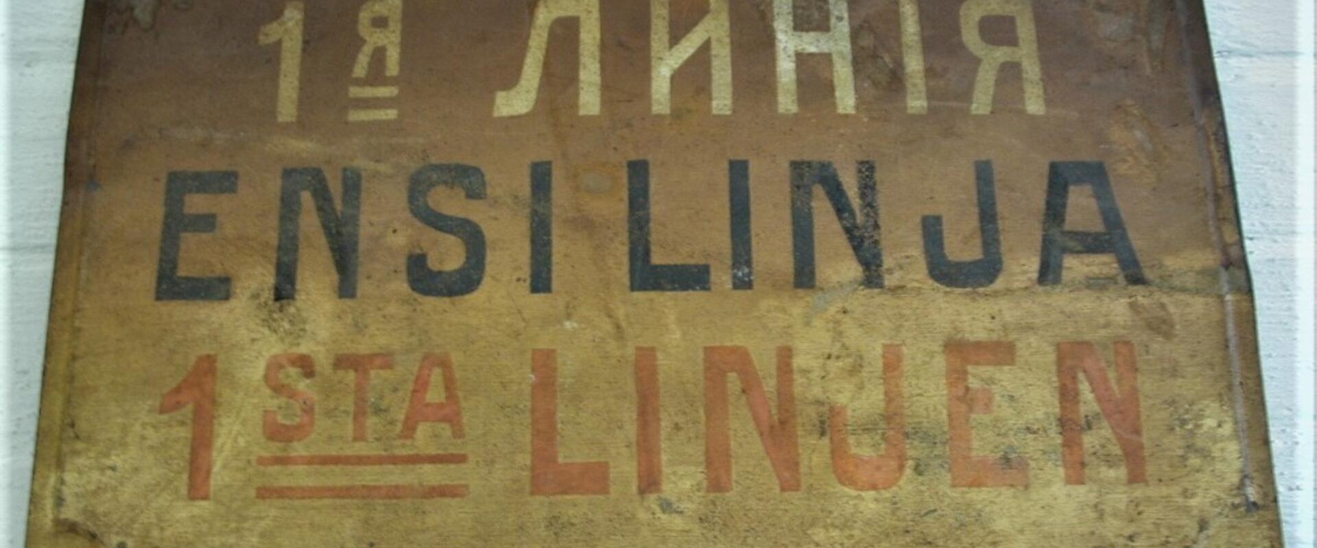Pohjaväri kellanruskea, kolmikielinen teksti: ENSI LINJA; 1. venäjä valkoisella, 2. suomi mustalla, 3. ruotsi punaisella värillä.