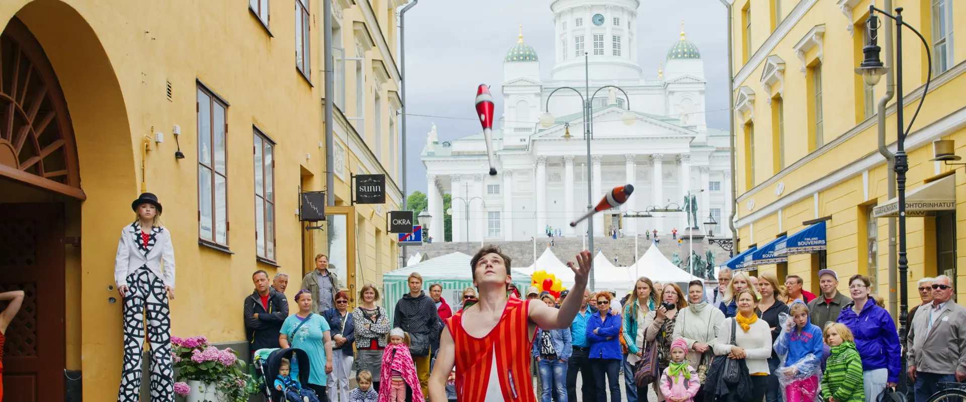 Etualalla jonglööri heittelee keiloja. Väkijoukko katselee taustalla, taka-alalla Tuomiokirkko.