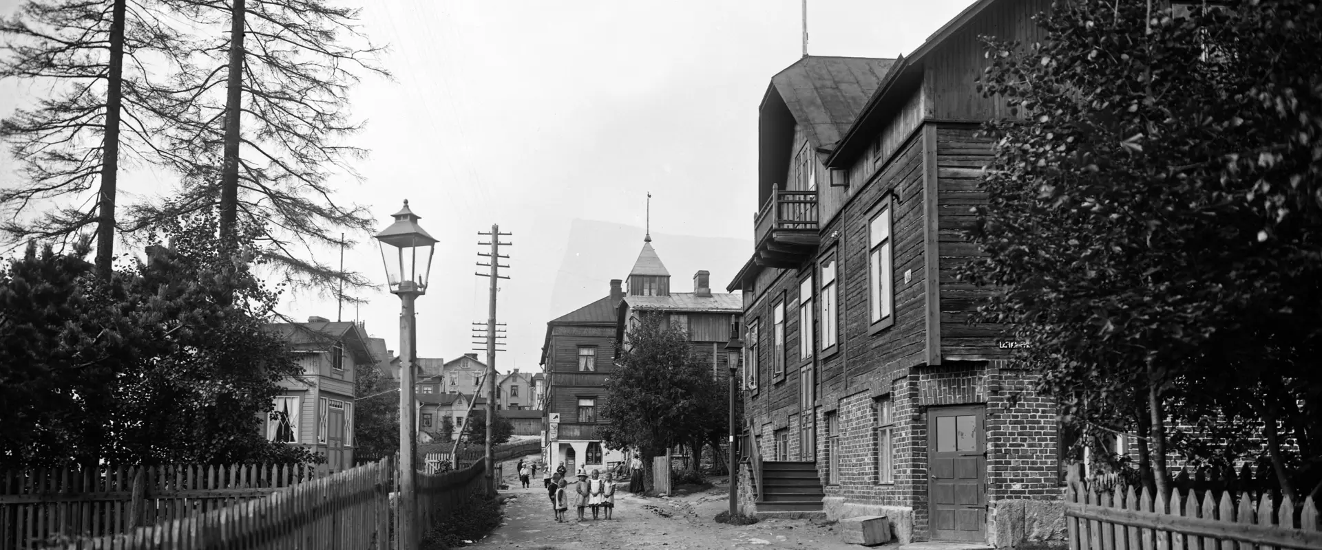 Katunäkymä Länsi-Pasilassa vuonna 1912. Kuvassa on työväen asuntoja suurempi puuhuvila. Hiekkatiellä kävelee muutama ihminen.