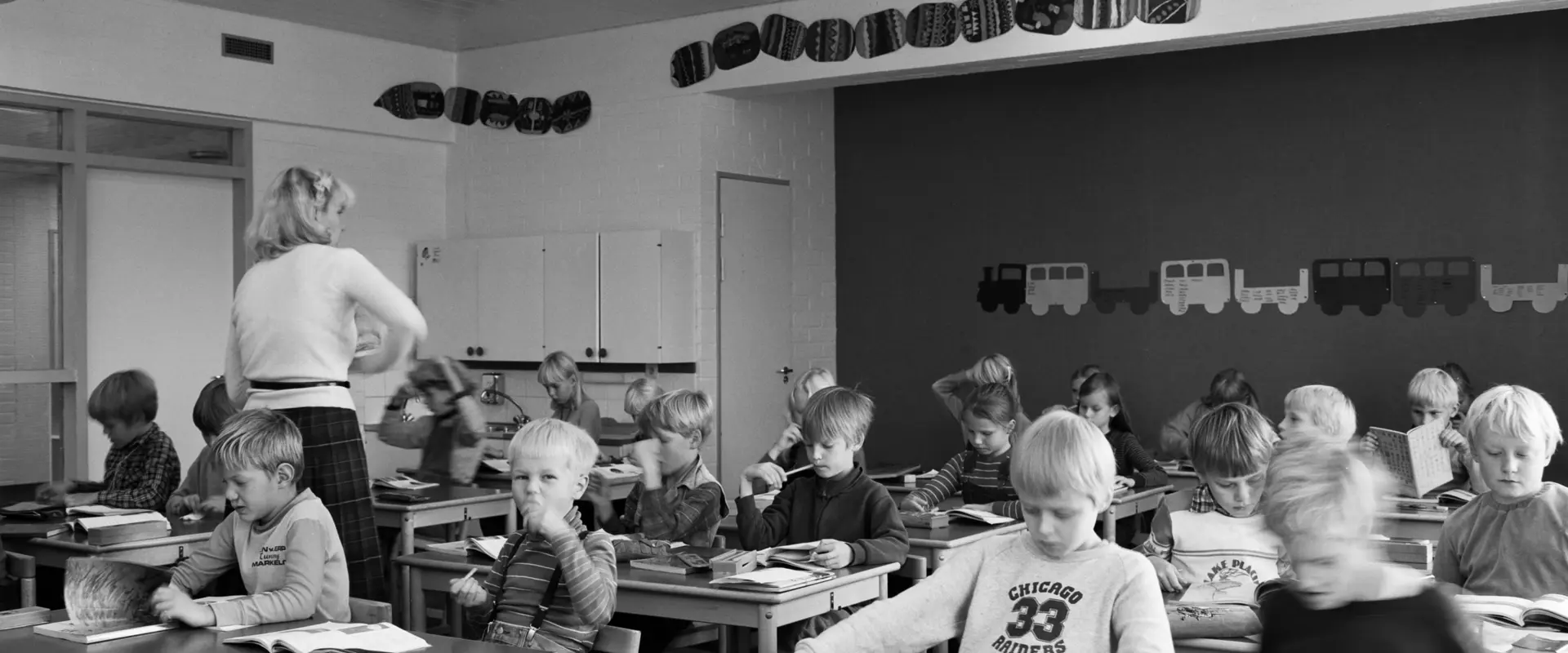 Alakoululaisia oppitunnilla luokassaan. Oppilaiden pöydillä on kirjapinoja, opettaja kiertää luokassa.
