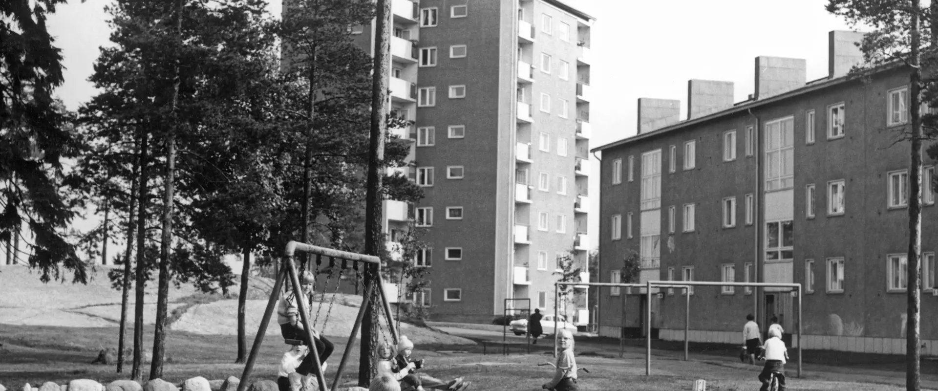 I förgrunden leker barn i en lekpark. En del av barnen är i sandlådan, några gungar och några har cyklar. I bakgrunden syns våningshus och träd. 