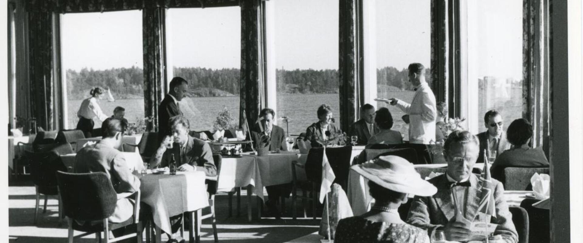 Pyöreä ravintolasali, jonka ikkunoista avautuu merinäköala. Salissa on tarjoilijoita ja asiakkaita valkoisella liinalla peitettyjen pöytien ääressä.
