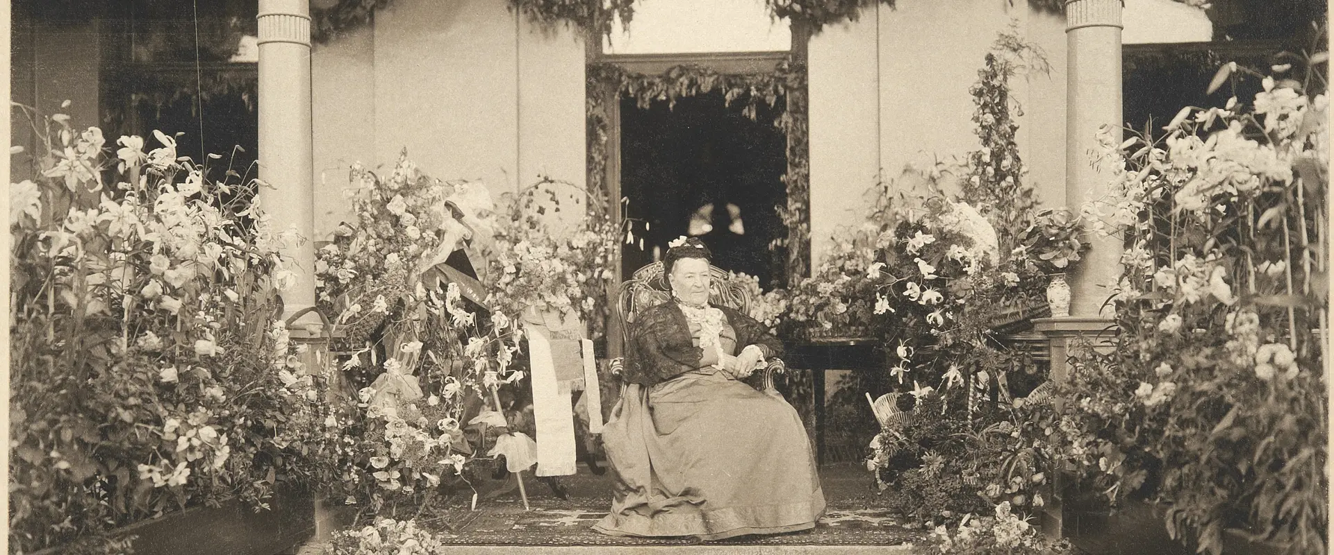 Aurora Karamzin fotograferad på sin 90-årsdag sittande framför ytterdörren till sin villa, omringad av blomplanteringar. En liten ljus hund sitter framför henne på trappan. 