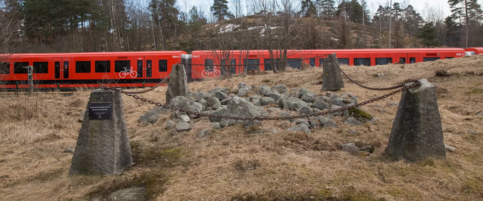 En gravhög från bronsåldern. Graven utgörs av en kvadratmeters område täckt med stenar av en fotbolls storlek. Graven är omringad av kättingar. I bakgrunden syns ett metrotåg.