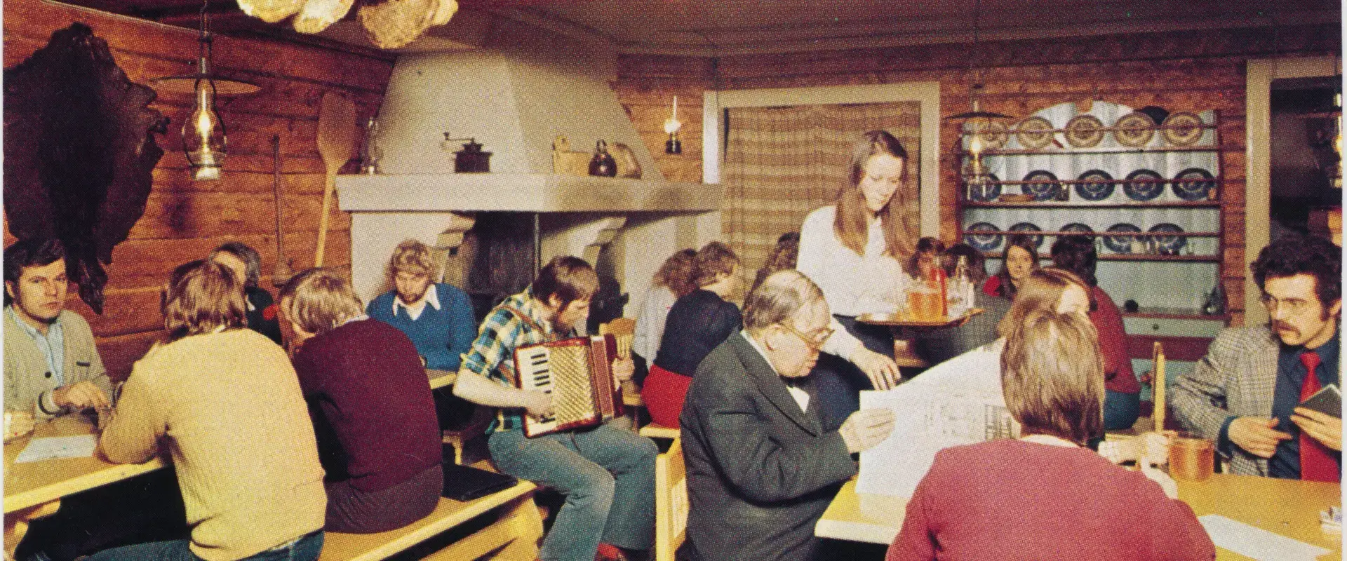 Pirtin tuvan tyyliin sisustettu ravintolatila, jossa ihmiset istuvat esimerkiksi lukemassa lehtiä. Mies soittaa haitaria, tarjoilija on tuomassa olutta ja limonadia tarjottimella. 