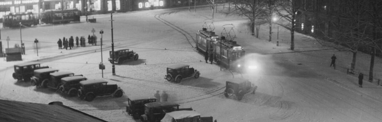 Katolta otetussa valokuvassa on luminen maisema Ruotsalaisen teatterin vierestä. Ihmisiä seisoo pysäkillä ja raitiovaunu on tulossa kääntöraiteelle. Autoja on pysäköity aukiolle.