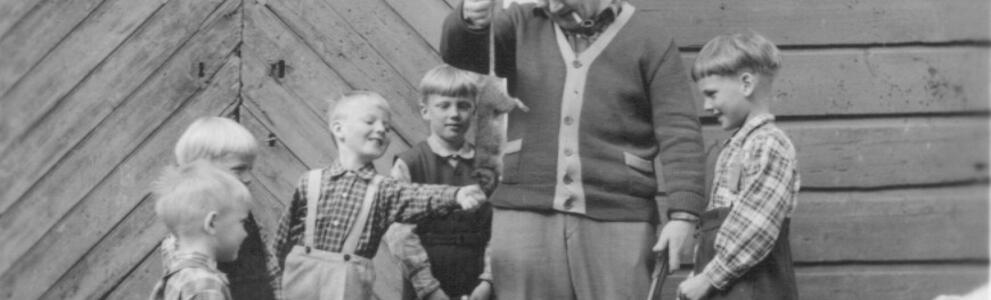 En man med pipa i munnen hänger en död råtta från svansen. En skara småpojkar pekar på djuret. 