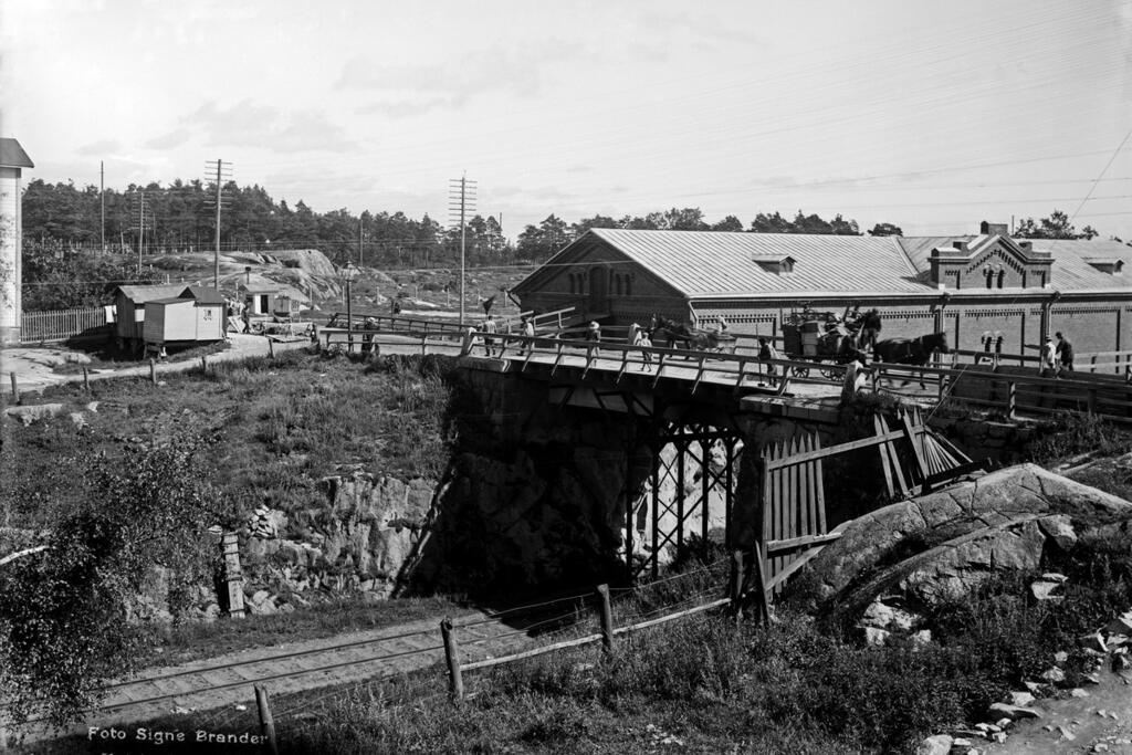 Trafik på Tavastvägens bro år 1909. Under bron syns järnvägen från Böle till Sörnäs hamn. Magasinsbyggnaden i rödtegel, som syns i bakgrunden, står fortfarande kvar, och på samma ställe finns en bro, om än större.  Foto: Helsingfors stadsmuseum / Signe Brander