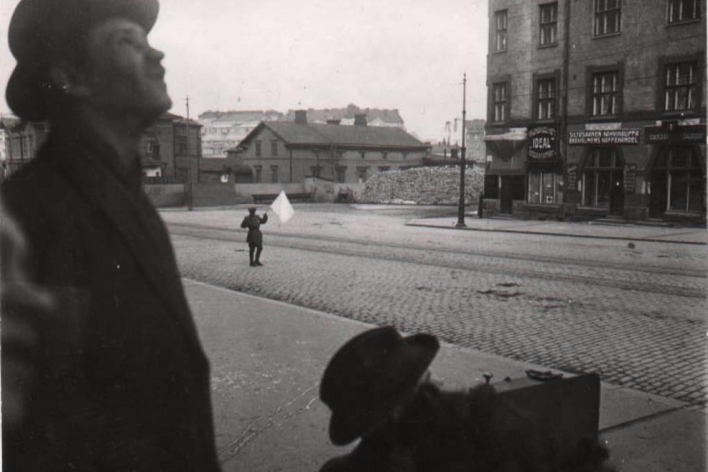 Punaisten antautuminen Siltasaarenkatu 5. Nuori mies kantaa valkoista antautumislippua autiolla kadulla. Kadun laidalla kaksi miestä katsovat oikealle yläviistoon. Toinen miehistä on kyykistynyt kuvaamaan tapahtumaa.

Viimeiset punaiset antautuivat Helsingissä 13.4. Helsingin valtauksen yhteydessä kuoli Sotatoimien aikana Helsingissä kuoli yli 400 henkilöä: saksalaisia 63, valkokaartilaisia 23, punaisia 320-340 ja siviilejä 10-20. Tuhansia punaisia jäi vangiksi. Kuvaaja: Tyyne Böök / Kansan Arkisto