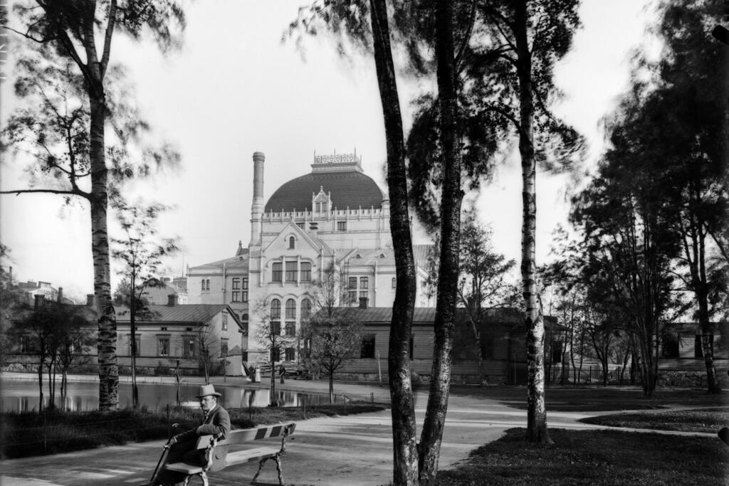 Svandammen, som låg i Kajsaniemiparken, var en kvarlämning av Gloviken, vars vatten hade letts till Tölöviken genom fördämning. Bilden är från år 1903, när Nationalteaterns byggnad som syns i bakgrunden var ny. De låga träbyggnaderna inhyste badhotellet Wilhelmsbad under åren 1864–1903. Foto: Helsingfors stadsmuseum / A. E. Rosenbröijer